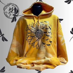 May include: A yellow hoodie with a large sunflower design on the front. The sunflower is black and white, with a cluster of black dragonflies to the right. The hoodie has a tie-dye effect and a hood.