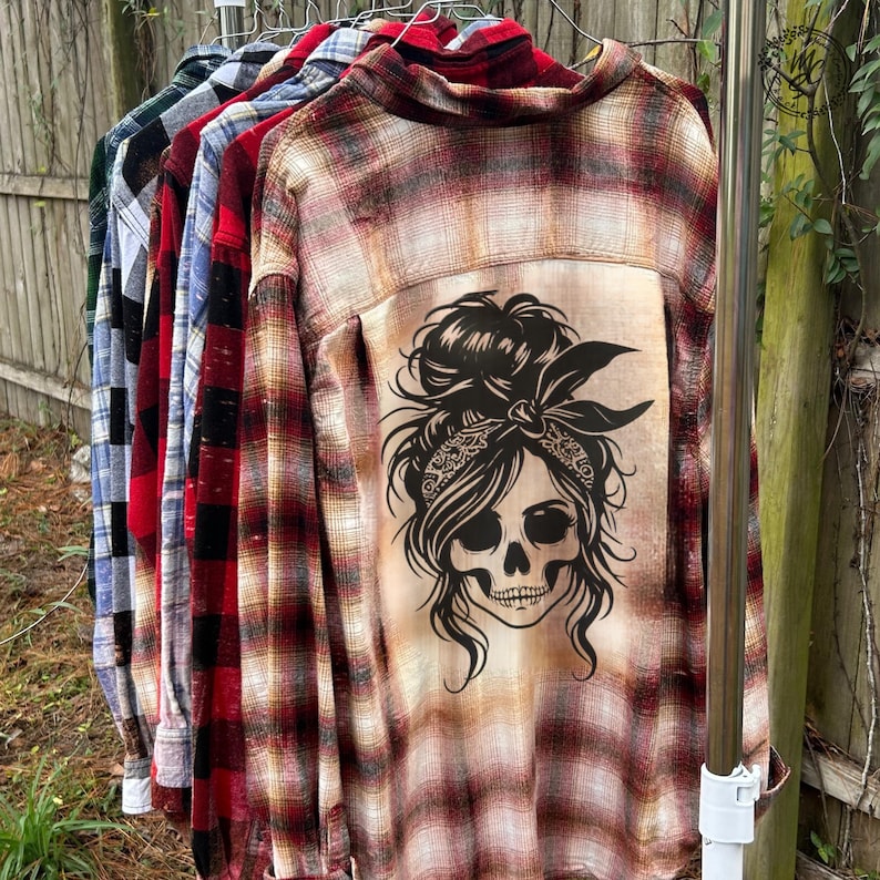 May include: A rack of plaid button-down shirts in various colors, including red, blue, and green. The shirts have a skull graphic on the back. The skull has a bandana and messy bun hairstyle.