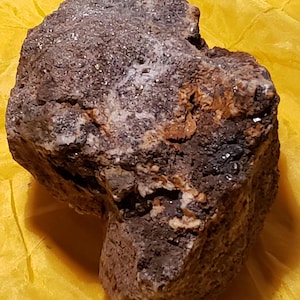 Könnte beinhalten: Ein unregelmäßig geformter Felsen mit einer dunkelbraunen und grauen Farbgebung. Die Oberfläche hat ein strukturiertes Aussehen mit kleinen, funkelnden Einschlüssen. Der Felsen liegt auf einem gelben Hintergrund.