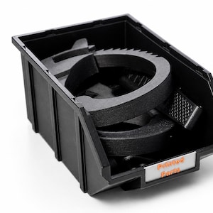 Peut inclure: Bac en plastique noir contenant plusieurs pièces noires, circulaires, imprimées en 3D. Le bac porte une étiquette orange avec l'inscription "Printed Parts". Les pièces sont probablement des composants d'un produit plus grand.