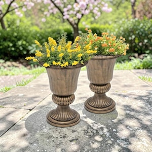 May include: Two bronze-colored pedestal planters with fluted bowls and bases. The left planter contains yellow flowers, while the right planter holds orange flowers. Both urn-shaped containers feature a textured finish and a wide rim.