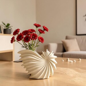 May include: A white ceramic vase shaped like a nautilus shell with a spiraling, ribbed texture. The vase holds a bouquet of vibrant red daisy-like flowers with green stems, creating a contrast between the smooth white form and the bright red petals.