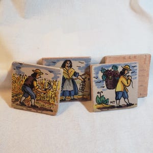 May include: Set of four square ceramic tiles with hand-painted scenes of people harvesting crops. The tiles feature figures in period clothing, with wheat fields, grapes, and a basket. The tiles are tan with blue and yellow accents.