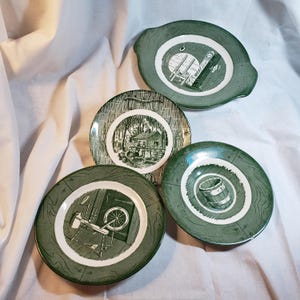 May include: Four green and white decorative plates with different illustrations. One plate has a handle. The illustrations include a spinning wheel, a cabin in the woods, a barrel, and a structure with a ladder.