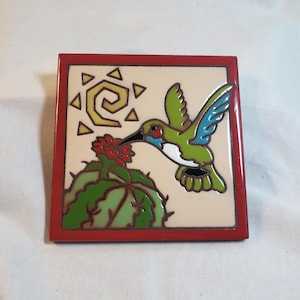 May include: Square ceramic tile with a red border featuring a hummingbird and cactus design. The hummingbird is green, blue, and white, with a long black beak, feeding from a red flower. A yellow sun is in the upper left corner.
