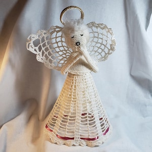 May include: A handmade crochet angel figurine in ivory with a gold halo and white hair. The angel has closed eyes and folded hands. The skirt of the angel is adorned with a thin, red ribbon. The wings are made of a delicate, open-weave crochet.