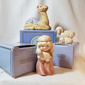 Può includere: Una collezione di statuine in ceramica raffiguranti un cammello, un pastorello con un agnello e pecore. Il cammello è marrone con una sella viola. Il pastorello indossa una veste rosa. Le pecore sono bianche.