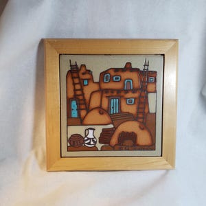 May include: A square ceramic tile in a light wood frame featuring a painted scene of brown adobe buildings with ladders, blue accents, and pottery in the foreground. The artwork includes the text '82 Teissadre' in the bottom right corner.