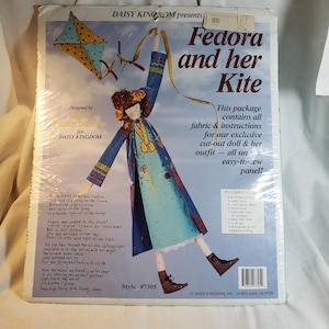 May include: A craft kit package titled "Fedora and her Kite" featuring a fabric doll design. The package includes instructions and fabric for a cut-out doll and outfit. The design includes a doll with a kite and text.