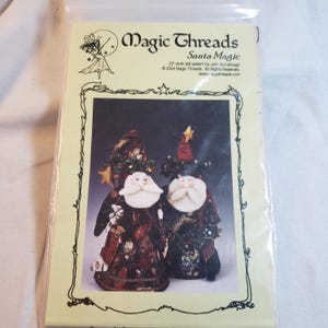 Op de afbeelding: Een pakket met een stoffen poppenpatroon getiteld "Santa Magic" van Magic Threads. Het patroon bevat afbeeldingen van twee kerstmanpoppen met witte baarden, gekleed in feestelijke outfits en hoeden. Het pakket is verzegeld in helder plastic.