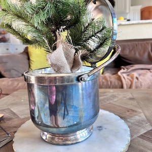 May include: A small, artificial Christmas tree in a silver ice bucket. The tree is wrapped in burlap and sits on a white marble coaster with a gold edge. The bucket has a handle and a lid that is partially open.