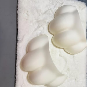 May include: Two off-white, cloud-shaped decorative pieces. The pieces have a smooth, matte finish and are arranged on a white, fluffy surface. The cloud shapes have rounded lobes, suggesting a soft, organic form. The lighting creates subtle shadows.