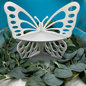 May include: A white butterfly-shaped decorative shelf with a circular platform. The butterfly wings have oval cutouts. The shelf is surrounded by green foliage against a blue background.