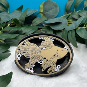 May include: A black oval coaster with a white and gold 3D design of a goldfish with bubbles. The coaster is on a white fuzzy surface.