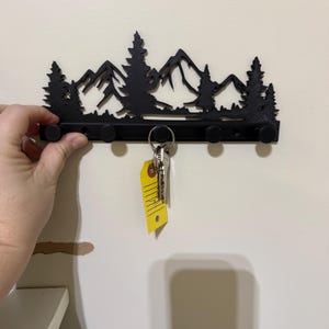 May include: Black metal key holder featuring a mountain and tree silhouette design. The key holder has five pegs and is holding a set of keys with a yellow tag. The key holder is mounted on a white wall.