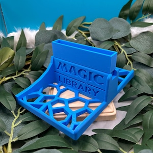 May include: A blue 3D printed Magic Library card holder with a geometric design. The holder has a slot for cards and a base for storing cards.
