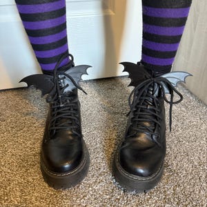 May include: A pair of black leather combat boots with thick soles, adorned with bat wing-shaped embellishments tied to the laces. The boots are paired with purple and black striped socks, creating a gothic or Halloween-themed aesthetic.