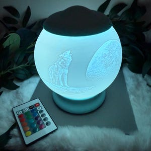 May include: A glowing, spherical lamp with a wolf howling at the moon design. The lamp emits a soft blue light and includes a remote control with color options. The lamp has a dark gray top and a light blue base.