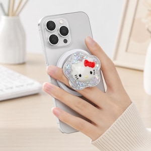 May include: A silver smartphone featuring a circular, flower-shaped phone grip attached to the back. The grip has a clear, glittery, iridescent finish and features a three-dimensional Hello Kitty character face with a bright red bow on its head.