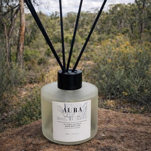 May include: Aura Australian Bush reed diffuser in a frosted glass bottle with black reeds. The label features the word "Aura" and the text "Australian Bush Reed Diffuser". The diffuser is set against a natural outdoor backdrop.