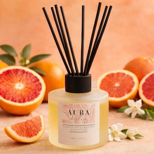 May include: Aura reed diffuser with black reeds, surrounded by blood oranges and grapefruit halves. The label reads "Blood Orange & Grapefruit Reed Diffuser". The background is a warm orange.