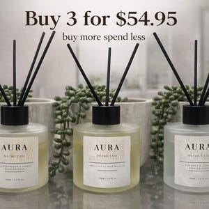 May include: Three glass reed diffusers with black reeds and black caps. Each diffuser has a label with the word "AURA" and the District Co. logo. The text "Buy 3 for $54.95" and "buy more spend less" are at the top.