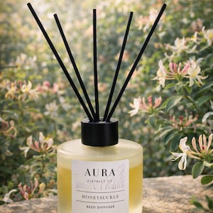 May include: Aura Honeysuckle reed diffuser with black reeds. The clear glass bottle contains a yellow liquid and has a black cap. The label reads "Aura District Co Honeysuckle Reed Diffuser 100ml / 3.4 fl oz."