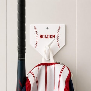 May include: White baseball home plate-shaped wall hook with the name "HOLDEN" in red. Red stitching detail and a white hook are visible. A baseball bat and a red and white striped bag hang below.