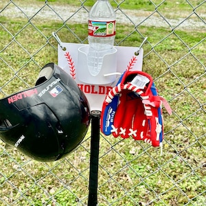 Pu&ograve; includere: Un set da baseball che include un casco nero, un guanto da baseball rosso e blu e una mazza nera. Una bottiglia d'acqua &egrave; in un supporto bianco attaccato a una recinzione a catena. &Egrave; presente anche un cartello con cuciture da baseball.