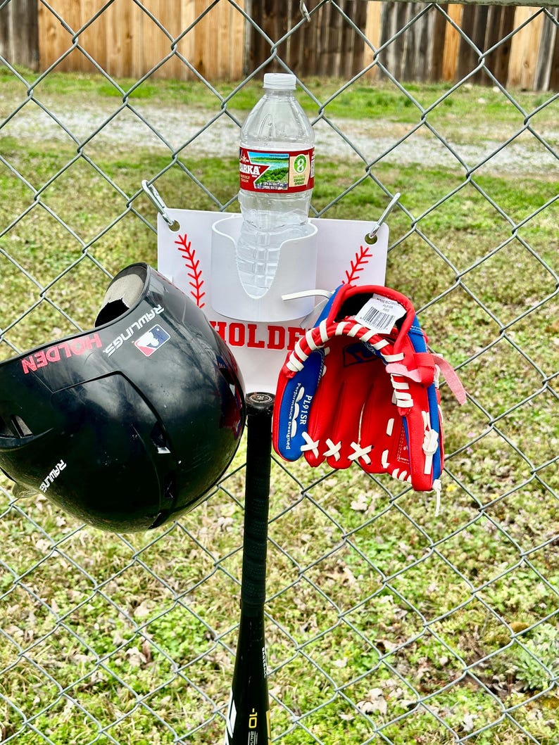 Pu&ograve; includere: Un set da baseball che include un casco nero, un guanto da baseball rosso e blu e una mazza nera. Una bottiglia d'acqua &egrave; in un supporto bianco attaccato a una recinzione a catena. &Egrave; presente anche un cartello con cuciture da baseball.