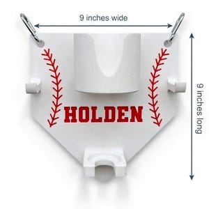 Pu&ograve; includere: Supporto a tema baseball bianco, largo e lungo 23 cm, con dettagli di cuciture rosse e il nome "HOLDEN" in lettere rosse. Presenta un portabicchieri centrale e due supporti laterali, con moschettoni argentati nella parte superiore.