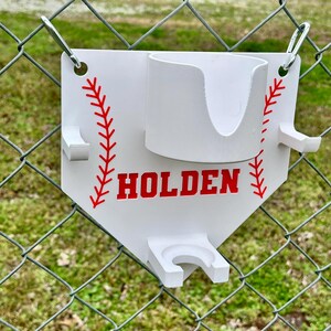 Pu&ograve; includere: Supporto bianco a tema baseball con dettagli di cuciture rosse e il nome "HOLDEN" in lettere rosse. Presenta un portabicchieri e due supporti aggiuntivi. Fissato a una recinzione a rete metallica con anelli metallici.