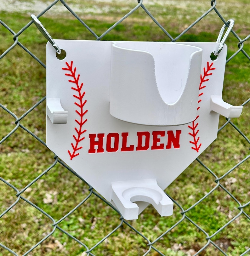Pu&ograve; includere: Supporto bianco a tema baseball con dettagli di cuciture rosse e il nome "HOLDEN" in lettere rosse. Presenta un portabicchieri e due supporti aggiuntivi. Fissato a una recinzione a rete metallica con anelli metallici.