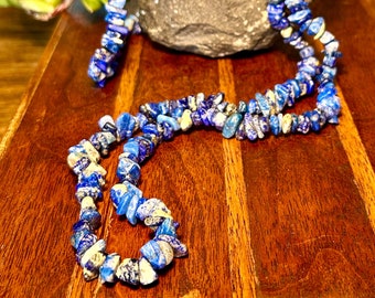 May include: A blue lapis lazuli necklace with irregular, chip-like beads. The necklace is displayed on a wooden surface, with a dark gray rock and greenery in the background. The beads have a mix of blue and white tones.