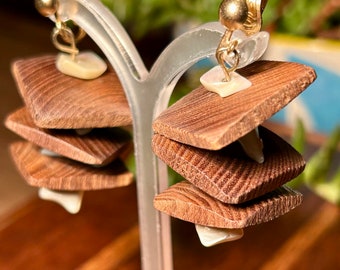 May include: Clip-on earrings featuring stacked, square wooden elements with a visible wood grain. Each square is separated by a small, white, shell-shaped accent. The earrings have gold-colored clips and are displayed on a clear earring stand.