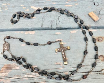 May include: A rosary featuring dark, oval-shaped beads linked by a metal chain. The piece includes a central metal medal depicting a figure and a crucifix pendant with a silver-toned corpus attached to a dark, rectangular cross frame.