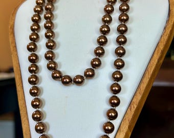 May include: A double strand necklace featuring lustrous, round, brown beads. The necklace is displayed on a white, angled stand with a wooden frame. The beads have a polished, reflective surface, creating a classic and elegant look.