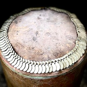 May include: A close-up of a silver necklace made of small, round, coin-shaped links. The necklace is lying on a brown, textured surface.