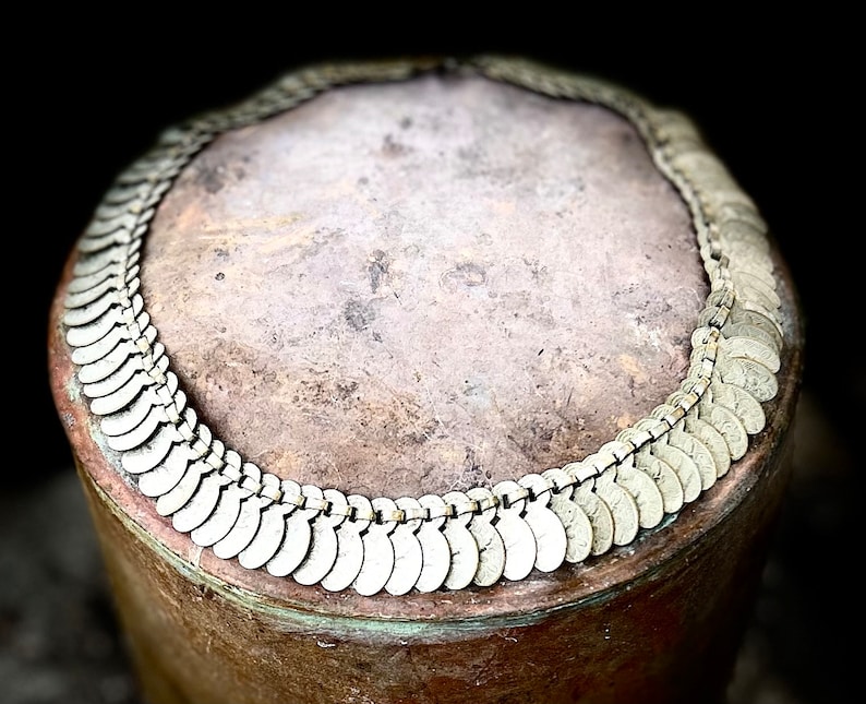 May include: A close-up of a silver necklace made of small, round, coin-shaped links. The necklace is lying on a brown, textured surface.
