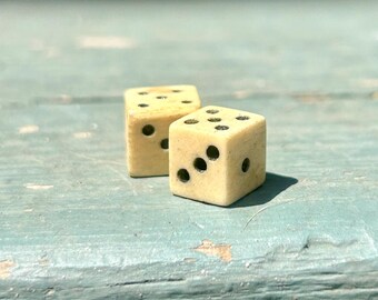 May include: Two vintage dice with black dots, each showing different numbers. The dice are a light beige color and appear to be made of bone or a similar material. They are resting on a weathered, light blue surface.