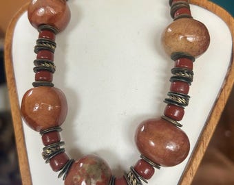 May include: A necklace with large, reddish-brown beads interspersed with smaller red beads and brass-colored spacers. The necklace is displayed on a white surface, showcasing its earthy tones and unique design.