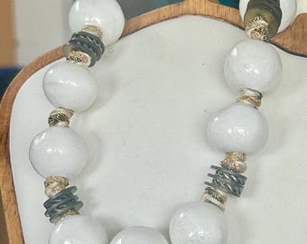 May include: A white beaded necklace with large, round beads. The necklace features dark gray and gold-colored accents between the white beads. The necklace is displayed on a white surface.