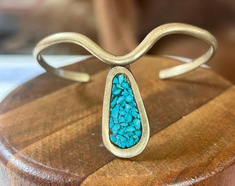 May include: A silver-toned cuff bracelet with a teardrop-shaped turquoise inlay. The bracelet has a curved design and is displayed on a wooden surface. The turquoise inlay is made of small, irregular pieces of bright blue stone.