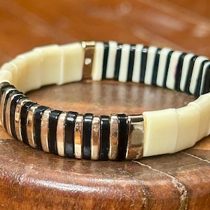 May include: A cream-colored and black striped bracelet with gold-colored accents. The bracelet is made of rectangular segments and is designed to be worn around the wrist. The bracelet is on a wooden surface.