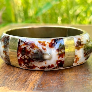 Puede incluir: Un brazalete de plata con incrustaciones de concha marrón y blanca y detalles dorados.