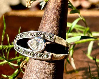 Könnte beinhalten: Ein silberner Ring mit einem birnenförmigen, klaren Stein, der in einem schwarzen Oval gefasst ist, das von kleinen schwarzen Steinen umgeben ist.