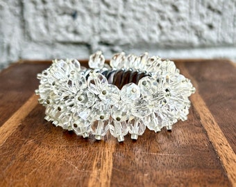 May include: A clear, beaded napkin ring with a silver-colored metal band. The beads are clustered around the ring, creating a sparkling, decorative accent. The napkin ring sits on a wooden surface.