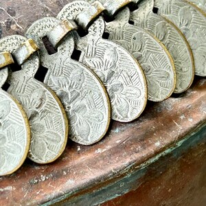 May include: A close-up of a silver metal chain with decorative, heart-shaped charms. The charms have a floral design and are attached to a copper surface.