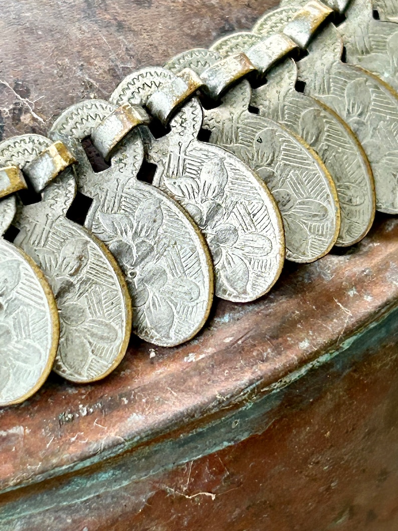 May include: A close-up of a silver metal chain with decorative, heart-shaped charms. The charms have a floral design and are attached to a copper surface.