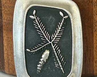 May include: A rectangular silver-toned belt buckle with a black center. The center features a detailed, stylized design of crossed branches with leaf-like elements. The buckle has a smooth, polished finish.